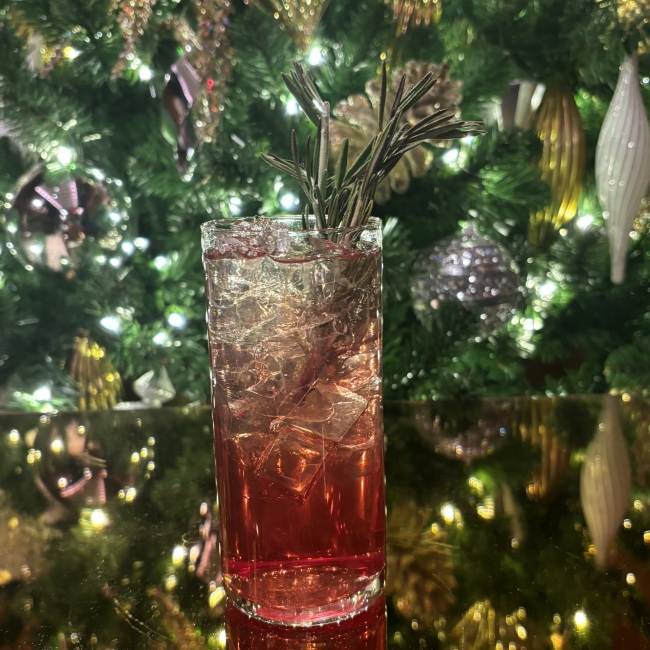 Cranberry colord cocktail in ornate highball glass in front of evergreen tree with lights